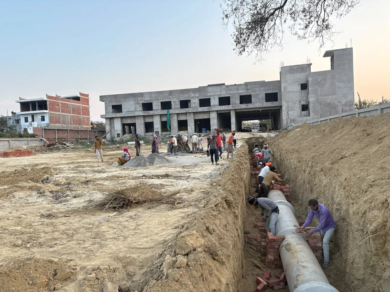 Crew laying a concrete pipeline in a trench beside a building under construction